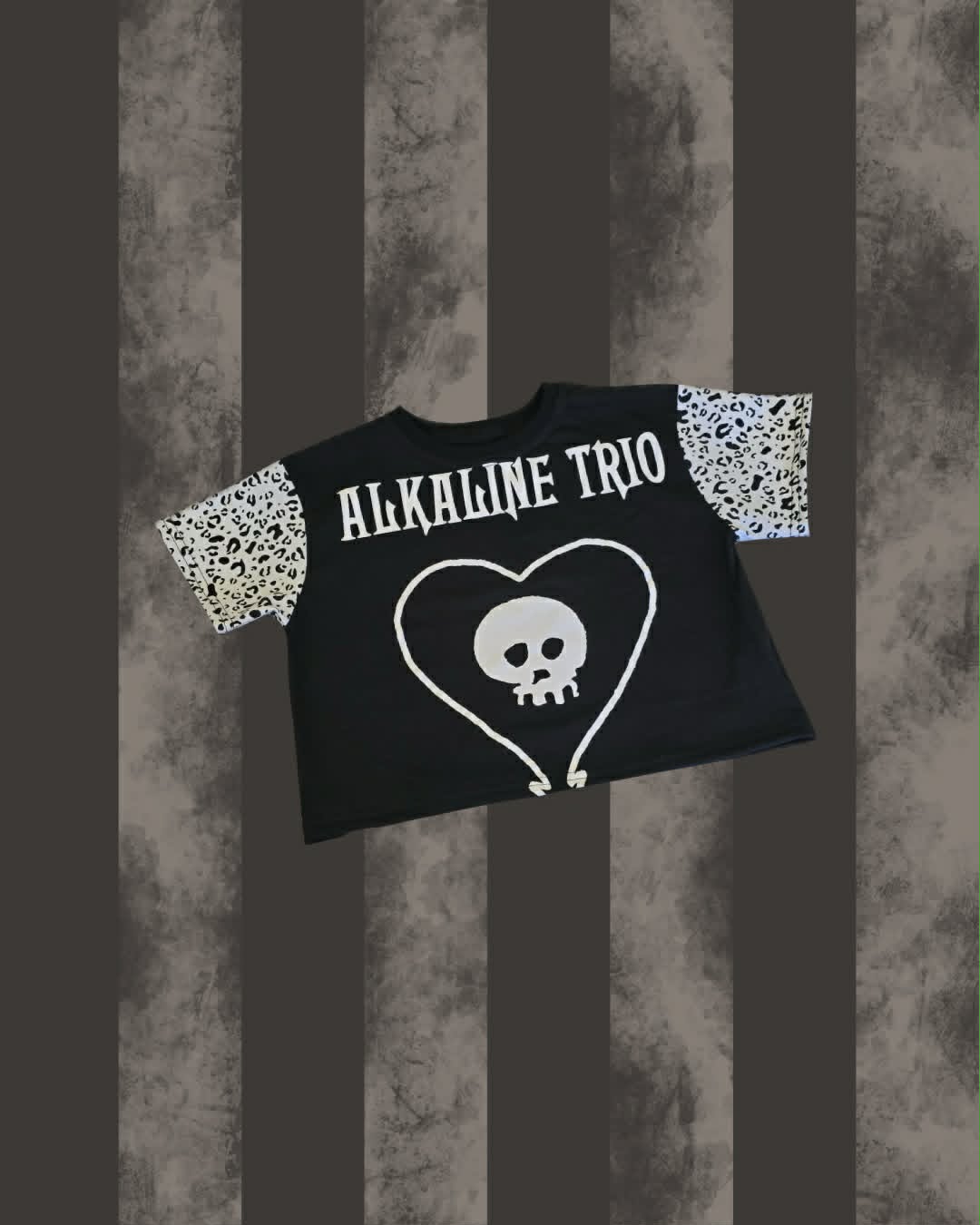 A black short-sleeve top featuring 'ALKALINE TRIO' in white text and a white skull-in-heart graphic on the front, with leopard print sleeves, shown against a dark gray and brown striped background.