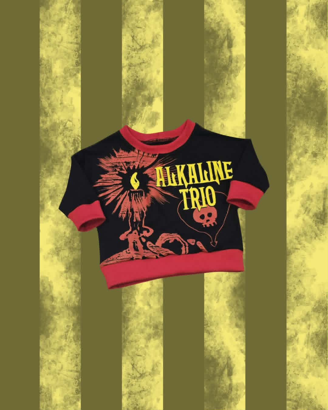 A sjumper featuring a black body with red collar, cuffs, and hem, printed with a red and yellow graphic of a skull, flame, and the text 'ALKALINE TRIO'. The shirt is displayed against a yellow and olive green vertically striped textured background.