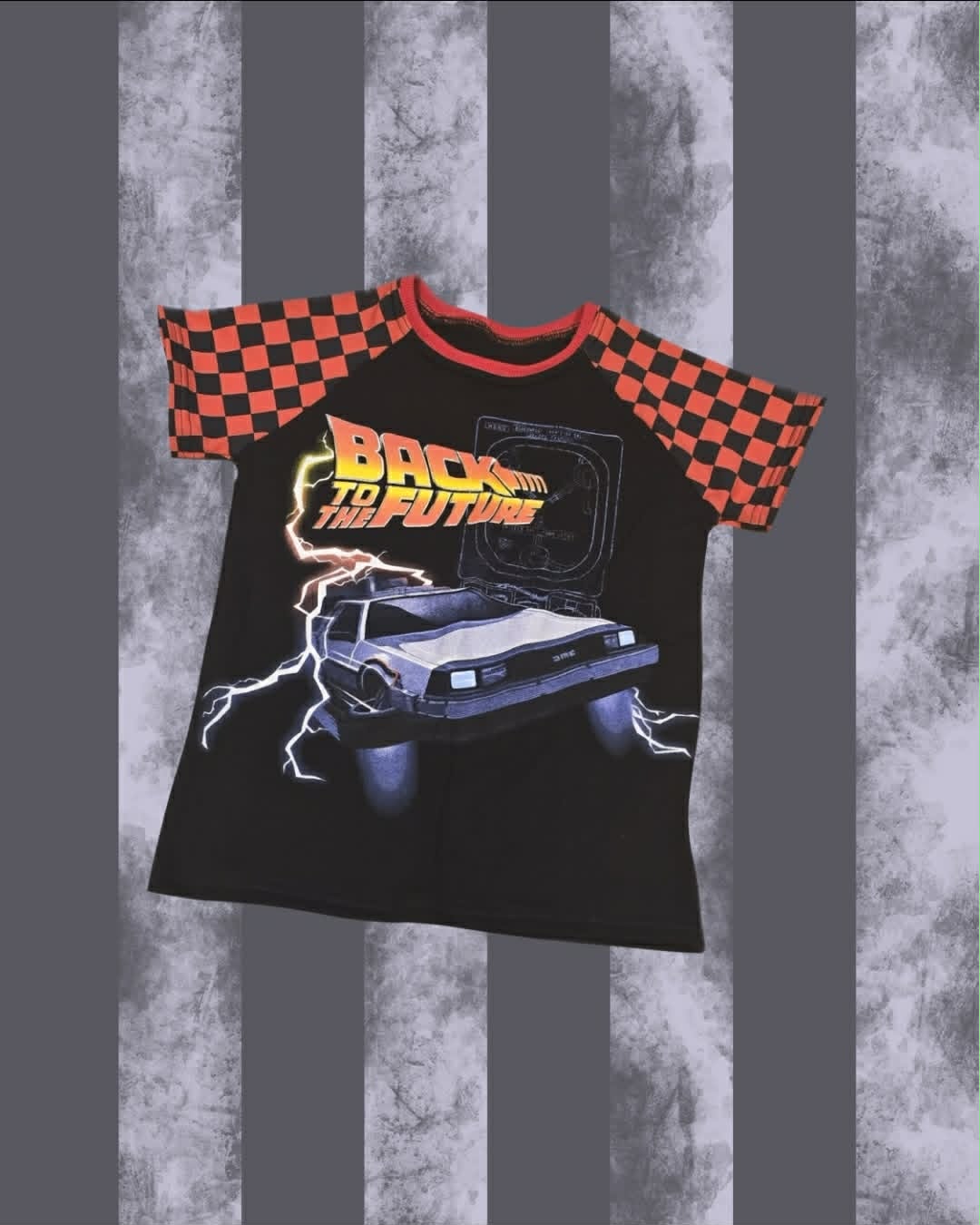 A black raglan t-shirt featuring a 'Back to the Future' DeLorean graphic on the front, with red and black checkered short sleeves and a red collar, displayed against a gray and white striped background.