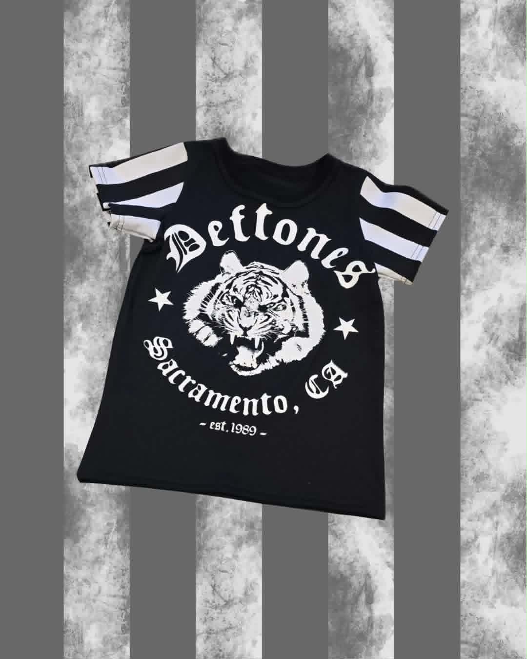 A black short-sleeve t-shirt with white graphic prints, featuring a tiger head in the center, the words 'Deftones' and 'Sacramento, Ca', and striped sleeves, shown against a gray and white textured background.