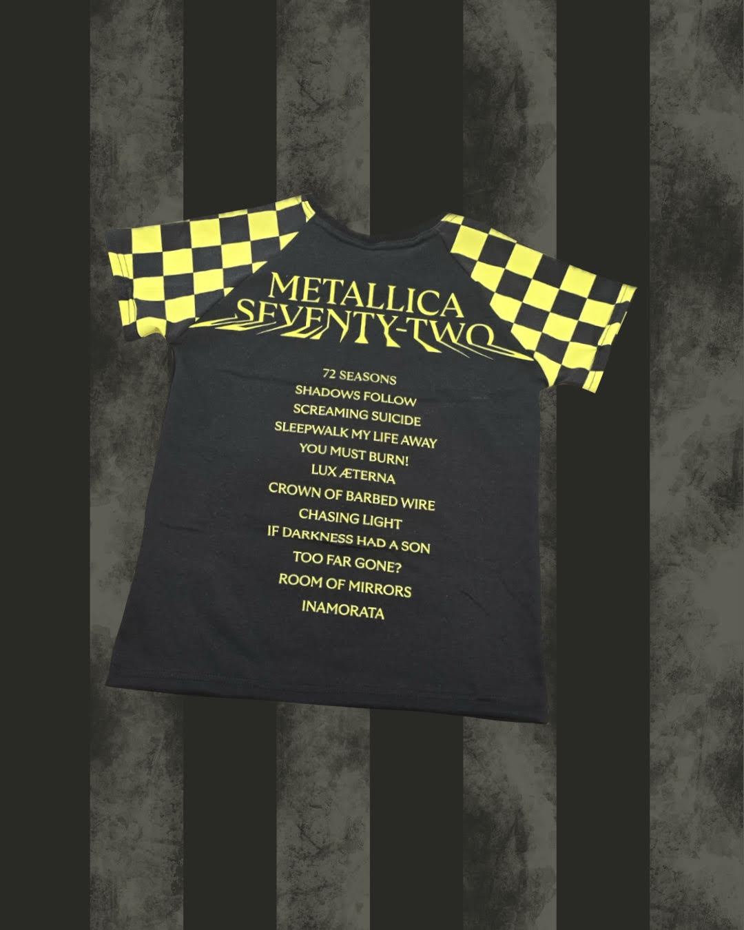 A black short-sleeve t-shirt with yellow and black checkered sleeves, featuring the text 'METALLICA SEVENTY-TWO' and a list of songs in yellow on the front, displayed against a dark gray and black striped background.