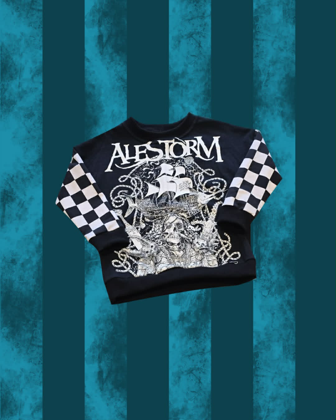 A black crewneck sweatshirt featuring a large white graphic print of a pirate ship and the word 'ALESTORM' across the chest, with black and white checkered patterns on the sleeves, displayed against a teal and dark teal vertical striped background.