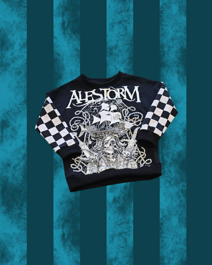 A black crewneck sweatshirt featuring a large white graphic print of a pirate ship and the word 'ALESTORM' across the chest, with black and white checkered patterns on the sleeves, displayed against a teal and dark teal vertical striped background.