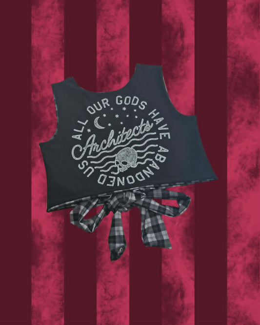 A sleeveless black top with a large white graphic reading 'All Our Gods Have Abandoned Us Architects' and a skull motif, tied at the waist with a plaid bow. The product is shown against a dark red vertically striped textured background.