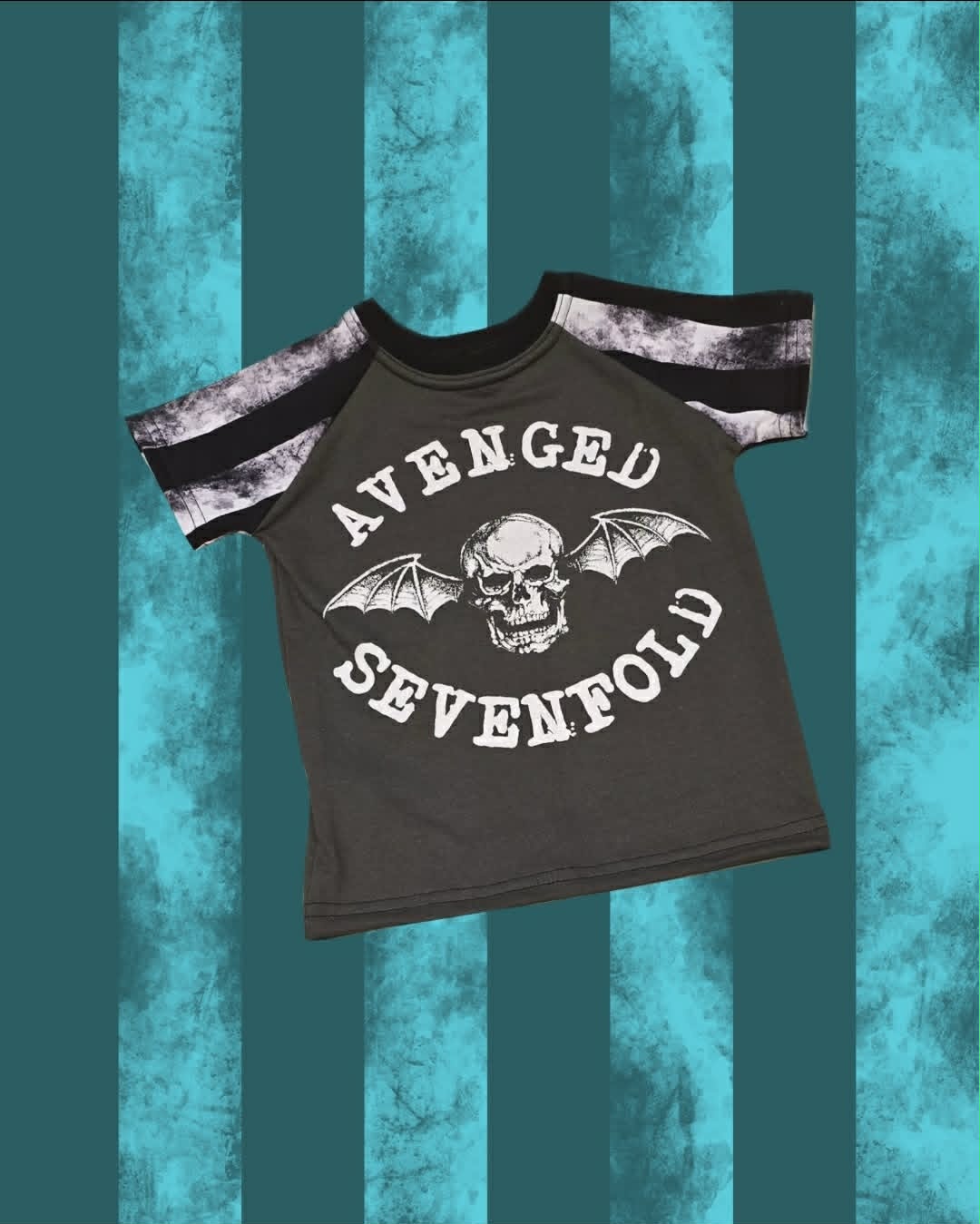 A dark gray short-sleeve t-shirt with a black and white tie-dye pattern on the sleeves, featuring a skull and bat graphic with the text 'AVENGED SEVENFOLD' on the front, set against a teal and black striped background.