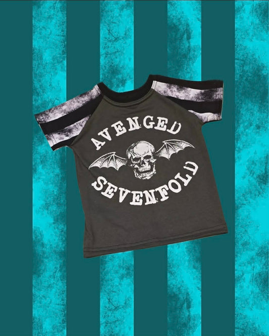 A dark gray short-sleeve t-shirt with a black and white tie-dye pattern on the sleeves, featuring a skull and bat graphic with the text 'AVENGED SEVENFOLD' on the front, set against a teal and black striped background.