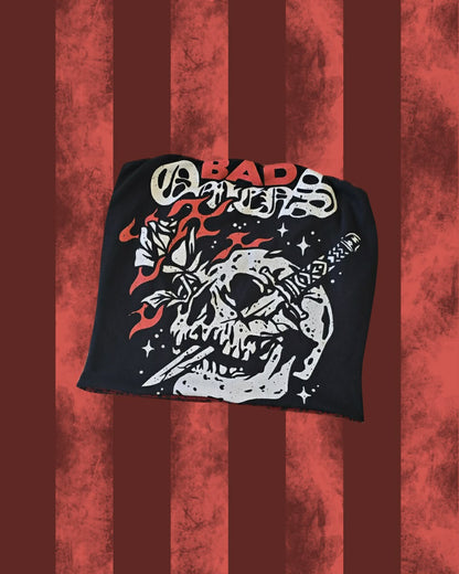 A black band t-shirt for 'Bad Omens' featuring a graphic of a skull holding a katana and a rose, with the band's name in stylized red, white, and black lettering. The shirt is displayed against a red and dark red vertically striped textured background.