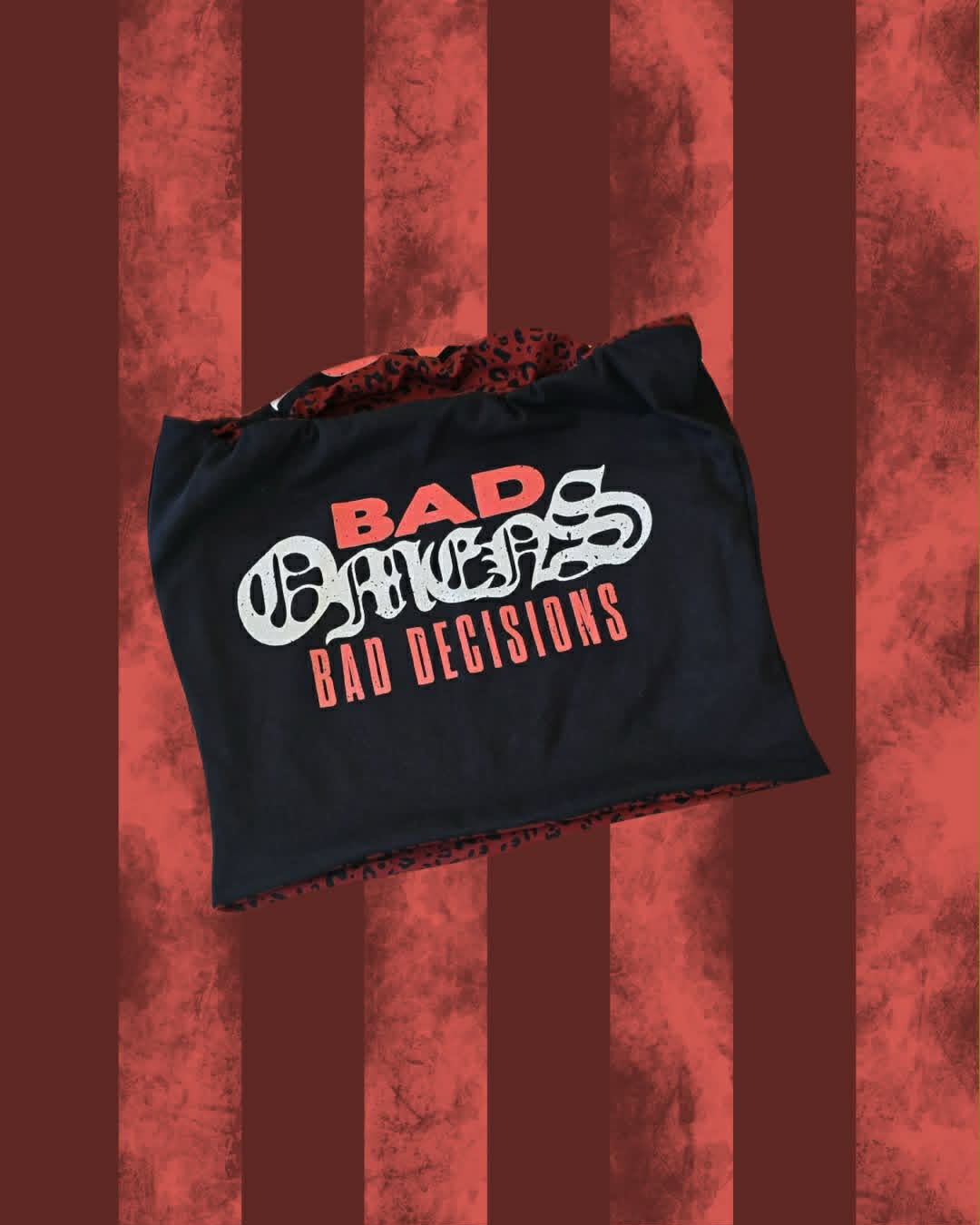 A black tote bag with a red and white graphic print reading 'BAD OMENS' and 'BAD DECISIONS' in red, shown against a red and dark red striped textured background.