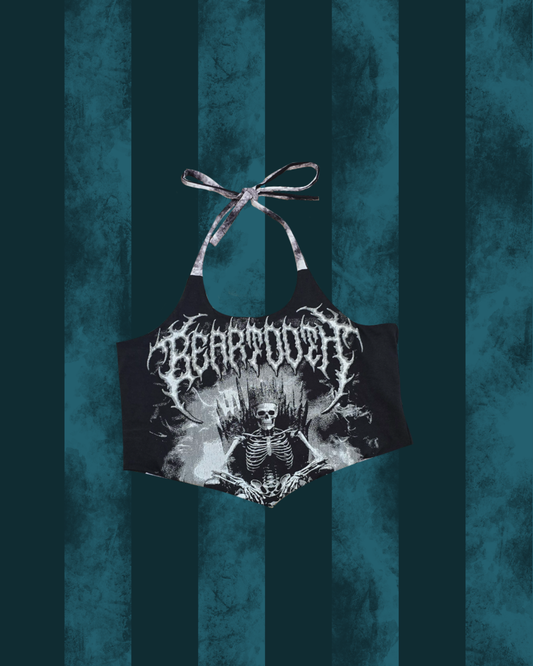 A black halter crop top featuring a white graphic print of a skeleton on a throne with the word 'BEARTOOTH' in a gothic font, displayed against a dark teal vertically striped background.