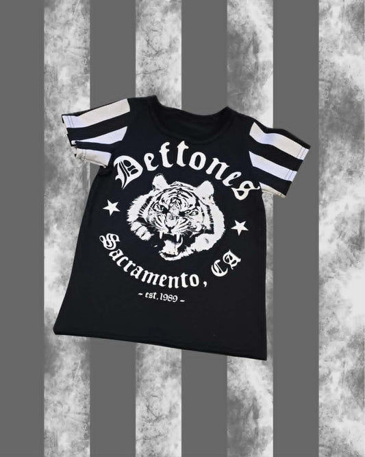 A black short-sleeve t-shirt with white graphic prints, featuring a tiger head in the center, the words 'Deftones' and 'Sacramento, Ca', and striped sleeves, shown against a gray and white textured background.