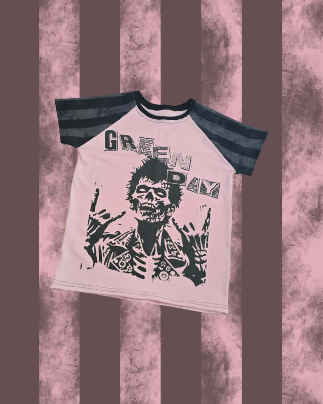 A Green Day 'Dilemma' raglan t-shirt with a light pink body, dark gray sleeves, and a black graphic print of the band's mascot on a pink and brown striped background.