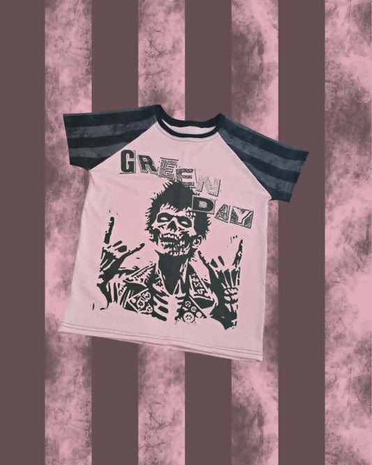 A Green Day 'Dilemma' raglan t-shirt with a light pink body, dark gray sleeves, and a black graphic print of the band's mascot on a pink and brown striped background.