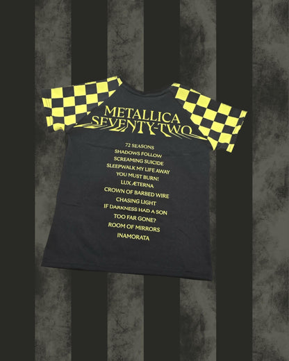 A black short-sleeve t-shirt with yellow and black checkered sleeves, featuring the text 'METALLICA SEVENTY-TWO' and a list of songs in yellow on the front, displayed against a dark gray and black striped background.