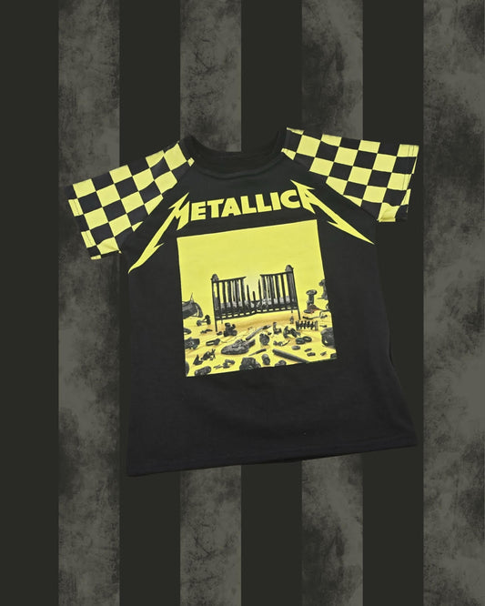 A black Metallica band t-shirt featuring the yellow 'Metallica' logo and 'The Iron Cage' album cover graphic on the front, with yellow and black checkered patterns on the shoulders, displayed against a dark gray and black striped background.