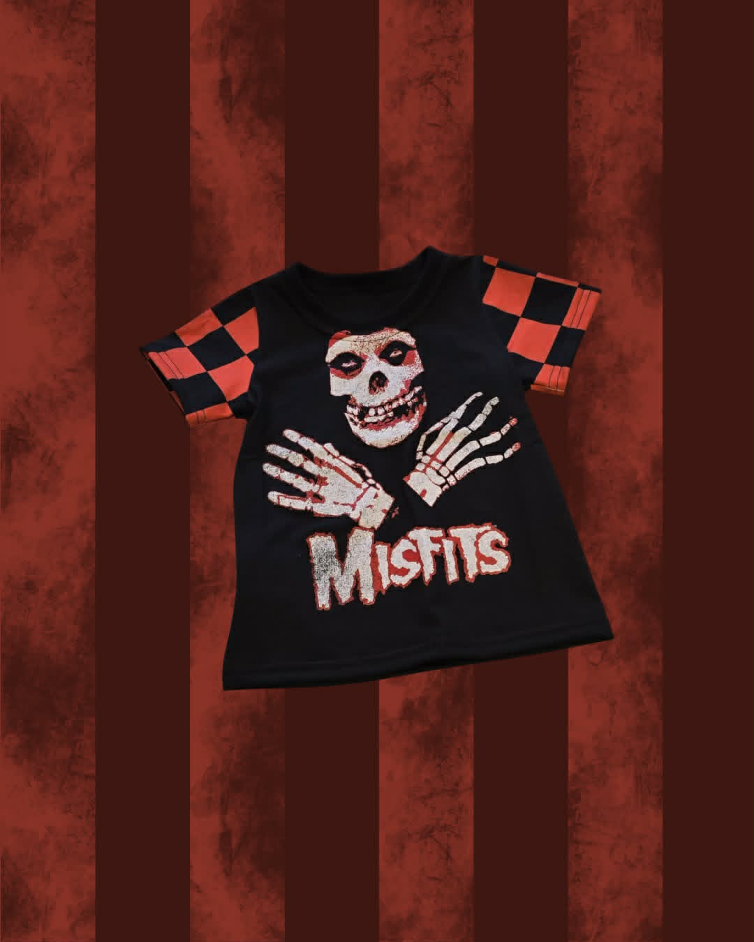 A black short-sleeve t-shirt with a horror-themed graphic featuring a skull, skeletal hands, and the text 'Misfits'. The t-shirt has orange and black checkered patterns on the sleeves.