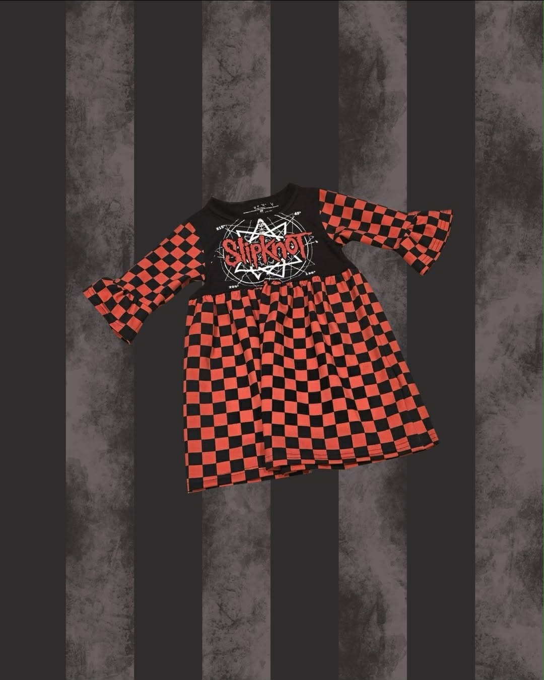 A children's long-sleeve dress with a black and red checkerboard pattern, featuring a solid black chest panel with a white and red 'Slipknot' band graphic. The dress is displayed against a dark gray and black vertically striped background.
