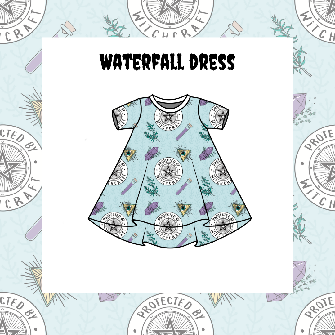 A kids' Waterfall Dress with short sleeves and a pattern of stars, planets, and waterfall-like designs on a blue background. The dress is presented on a dress form, which is protected by a star-shaped shield in the background.
