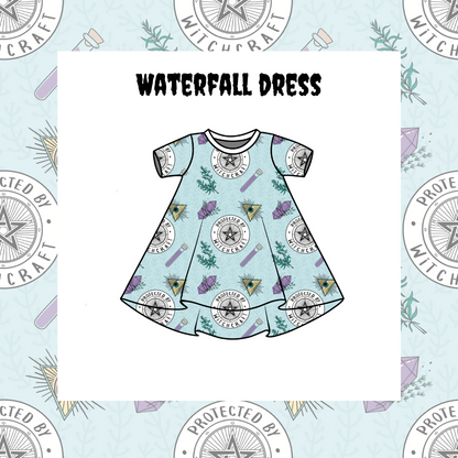 A kids' Waterfall Dress with short sleeves and a pattern of stars, planets, and waterfall-like designs on a blue background. The dress is presented on a dress form, which is protected by a star-shaped shield in the background.
