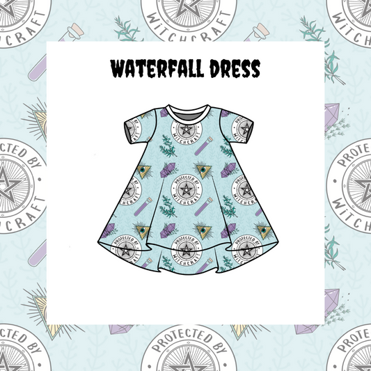 A kids' Waterfall Dress with short sleeves and a pattern of stars, planets, and waterfall-like designs on a blue background. The dress is presented on a dress form, which is protected by a star-shaped shield in the background.